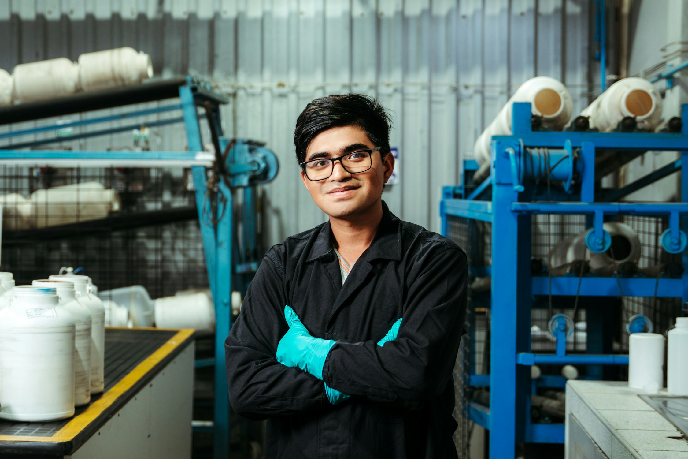 A man standing with his arms crossed on the floor of the plant he runs, machinery and stock behind him