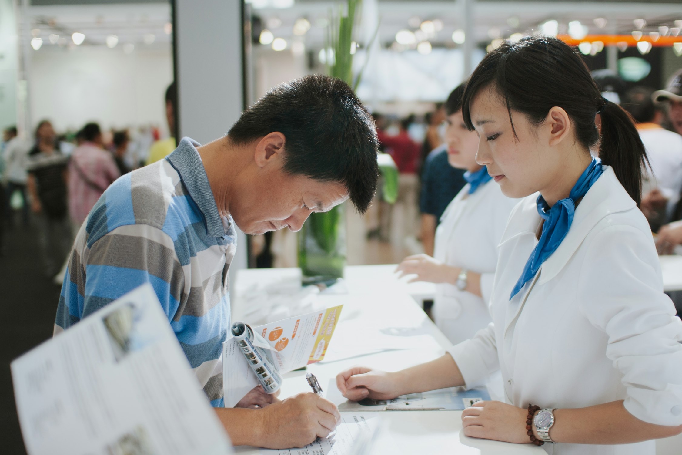 A man filling in a document at a trade stand counter spread with product literature, an attendant beside him
